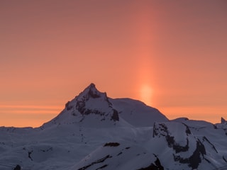 Verschneiter Berg, rötlicher Himmel mit hellerem, vertikalem Lichtstrahl.