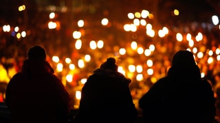 Schwarze Umrisse von drei Menschen, die auf ein Kerzenmeer blicken.