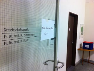 Ein Eingang zu einem Arztbüro in einem Spital. Auf der Türe ist ein Schild angebracht, auf dem "Maske / Garderobe" zu lesen ist.