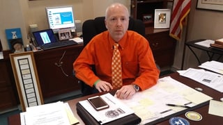 Ein Mann in orangem Hemd sitzt an einem Schreibtisch.
