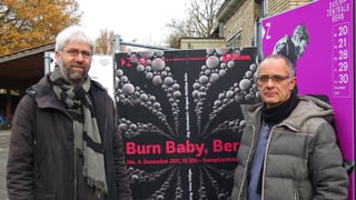 Zwi Männer stehen vor einem Plakat.