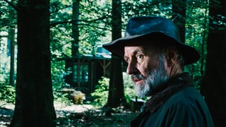 Ein Mann mit Hut steht im Wald vor einer Hütte