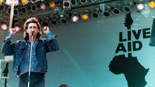 Ein Mann mit mittellangen Haaren und Jeansjacke auf der Bühne mit dem Logo Live Aid. Ein Mann mit mittellangen Haaren und Jeansjacke auf der Bühne mit dem Logo Live Aid.