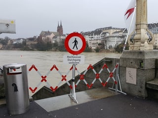 Vor einer Treppe steht rotweisse Schranke mit eine Verbotsschild für Fussgänger. Im Hintergrund sieht man den brauen Rhein und das Basler Münster.