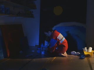 Die Künstlerin knieht im Dunklen am Boden, neben ihr Bilder und Maluntensilien.