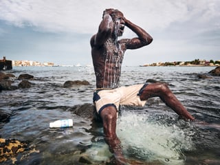 Mann sitzt auf einem Stein im Meer und giesst sich Milch über den Kopf.