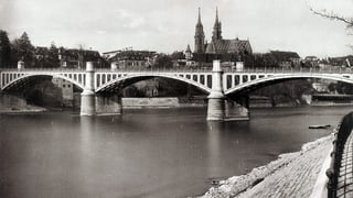 Schwarz-Weiss-Fotografie mit einer Brücke, die über einen Fluss führt. Dahinter die zwei Türme des Münsters.