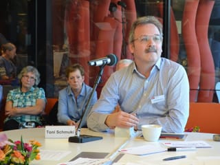 Toni Schmucki vor dem Mikrofon, hinter ihm sitzen zwei Frauen im Publikum.