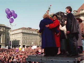 Dreifuss und Brunner stehen auf einem Podium vor der Menschenmenge auf dem Bundesplatz. Dreifuss hält eine rote Rose hoch.