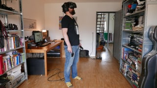 Eine Person steht in einem Wohnzimmer, trägt eine VR-Brille und hält Controller in der Hand.