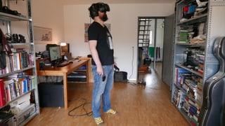 Eine Person steht in einem Wohnzimmer, trägt eine VR-Brille und hält Controller in der Hand. Eine Person steht in einem Wohnzimmer, trägt eine VR-Brille und hält Controller in der Hand.