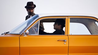Ein Mann in einem orangen Auto. Neben dem Auto geht ein Mann mit Hut.