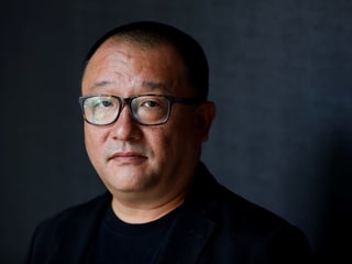 Nahaufnahme: Ein Mann mit asiatischer Herkunft schaut in die Kamera. Er hat kurze, schwarze Haare, trägt eine schwarze Hornbrille, ein schwarzes T-Shirt und darüber ein dunkles, samitges Jakett. Nahaufnahme: Ein Mann mit asiatischer Herkunft schaut in die Kamera. Er hat kurze, schwarze Haare, trägt eine schwarze Hornbrille, ein schwarzes T-Shirt und darüber ein dunkles, samitges Jakett.