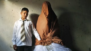 Ein Junge mit Hemd und Krawatte und ein vollverschleiertes Mädchen in einer weissen Hochzeitsrobe stehen nebeneinander.