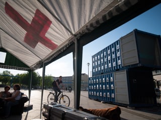 Blick aus einem weissen Zelt mit rotem Kreuz auf die blauen Schiffscontainer.
