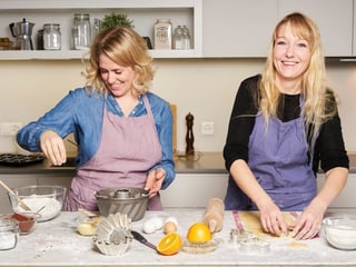 Katharina Rippstein und Claudia Boutellier stehen lachend in der Küche und backen.