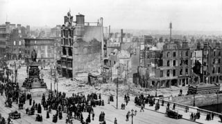 Schwarzweiss Aufnahme, Menschen im Vordergrund, im Hintergrund zerstörte Gebäude