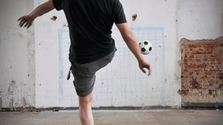 Ein junger Mann kickt einen Fussball in Richtung einer Wand, auf die mit blauer Kreide ein Tor gezeichnet ist.