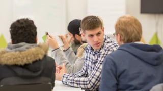 Ein Mann mit Karohemd sitzt vor einer Pinnwand mit Fotos, ihm gegenüber zwei Frauen mit Kopftuch. Ein Mann mit Karohemd sitzt vor einer Pinnwand mit Fotos, ihm gegenüber zwei Frauen mit Kopftuch.