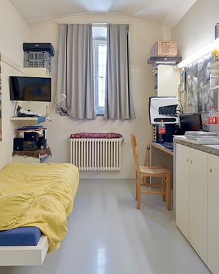 Eine Gefängniszelle: ein Bett, ein Schreibtisch, ein TV und ein Fenster.