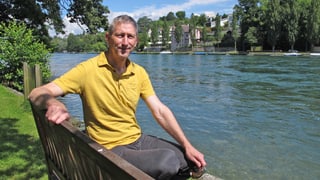 Ein Mann mit gelbem T-Shirt sitzt auf einer Bank neben einem Fluss.