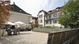 Blick auf einen Dorfplatz mit Tischen und Stühlen und historischen Gebäuden im Hintergrund. Blick auf einen Dorfplatz mit Tischen und Stühlen und historischen Gebäuden im Hintergrund.