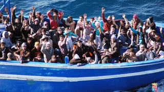 Ein volles Boot auf dem Meer vor Lampedusa, die Passagiere winken. Einer hält ein kleines Kind hoch.
