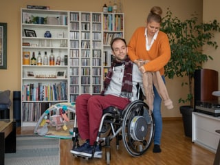 Eine stehende Frau zieht einem Mann im Rollstuhl die Jacke an. Eine stehende Frau zieht einem Mann im Rollstuhl die Jacke an.
