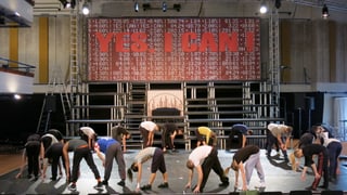 Tänzerinnen und Tänzer auf einer grossen Bühne, alle bücken sich nach vorne. Auf einem grossen Bildschirm über ihnen steht "yes, I can"