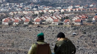 Zwei Soldaten von hinten stehen leicht erhöht und schauen auf die Siedlung mit Dutzenden gleichen Häusern.