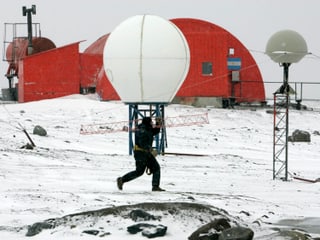 Ein Mann trägt eine Antenne vor einer Antarktisstation.