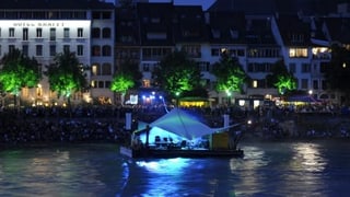 Das bläulich beleuchtete Floss schaukelt auf dem Rhein, von der Grossbasler Seite her fotografiert.