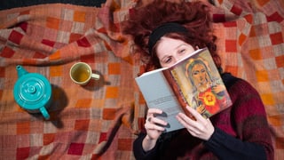 Eine Frau mit rotem Haar liegt auf einer Decke und liest ein Buch.