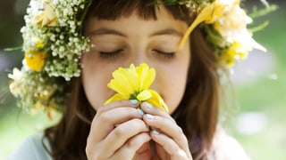 Ein Mädchen mit Blumenkranz auf dem Kopf riecht an einer gelben Blume. 