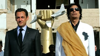 Links steht Nicolas Sarkozy, rechts Gaddafi, in der Mitte eine goldene Hand, die ein amerikanisches Flugzeug zerquetscht.