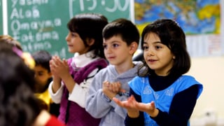 Drei Kinder stehen in einem Schulzimmer in einer Reihe, die Handflächen gegen oben gerichtet. 