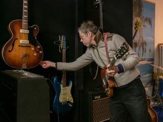 Mann hält eine Gitarre und greift mit der freien Hand nach einer zweiten Gitarre die an der Wand hängt Mann hält eine Gitarre und greift mit der freien Hand nach einer zweiten Gitarre die an der Wand hängt