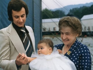 Eine alte Dame mit einem Baby im Taufkleid im Arm, daneben ein Mann im Anzug.