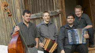 Die vier Musiker mit ihren Instrumenten sitzend und stehend vor einer Holzhütte.