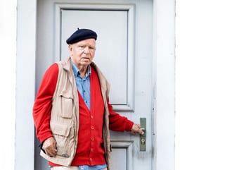 Ein Mann mit Béret steht vor einer Haustüre, die Hand an der Türklinke.