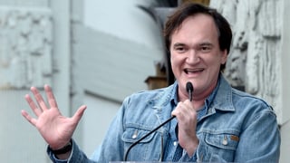 Quentin Tarantino vor einer Statue. Er spricht ins Mikrofon und hebt seine rechte Hand.