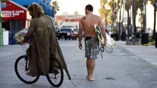 Ein Obdachloser fährt Rad. Neben ihm geht ein Surfer.