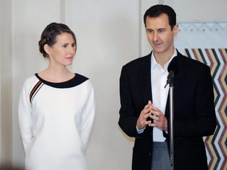 Assad redet in ein Mikrophon, seine Frau steht neben ihm