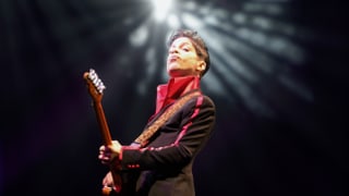 Prince spielt Gitarre, hinter seinem Kopf leuchtet ein Scheinwerfer.
