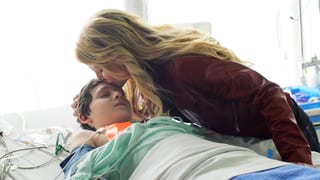 Eine junge Frau beugt sich in einem Spital über einen jungen, der mit geschlossenen Augen auf dem Bett liegt, und küsst ihn auf die Stirn.