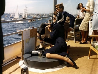 Ein Mann und eine Frau mit Sonnenbrille sitzen auf Deck eines Bootes und schauen einem Segelwettkampf zu.