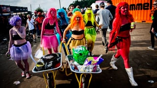 Sechs Frauen in neonfarbigen Kleidern und Schubkarren
