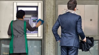 Weibliche Putzkraft reinigt einen Bankomaten, daneben ein Mann in Anzug (beide von hinten)