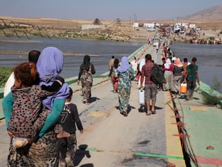 Frauen, Kinder und Männer gehen zu Fuss über eine Flussbrücke.