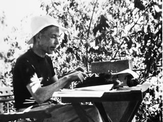 Friedrich Glauser, mit Hut und Zigarette im Mund draussen sitzend, auf einer Schreibmaschine schreibend.